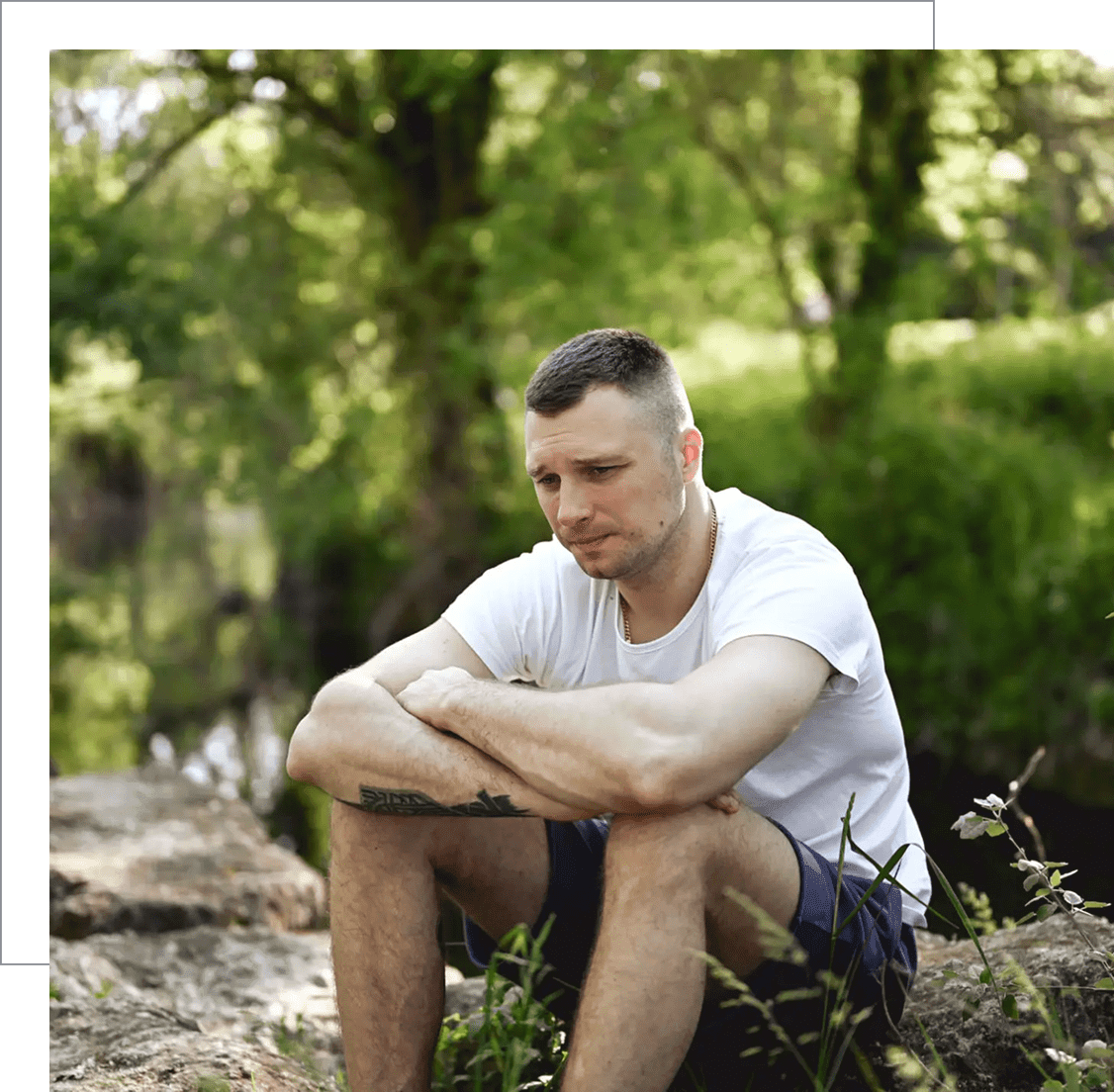 A man in a white t-shirt sitting thoughtfully outdoors in a forest.