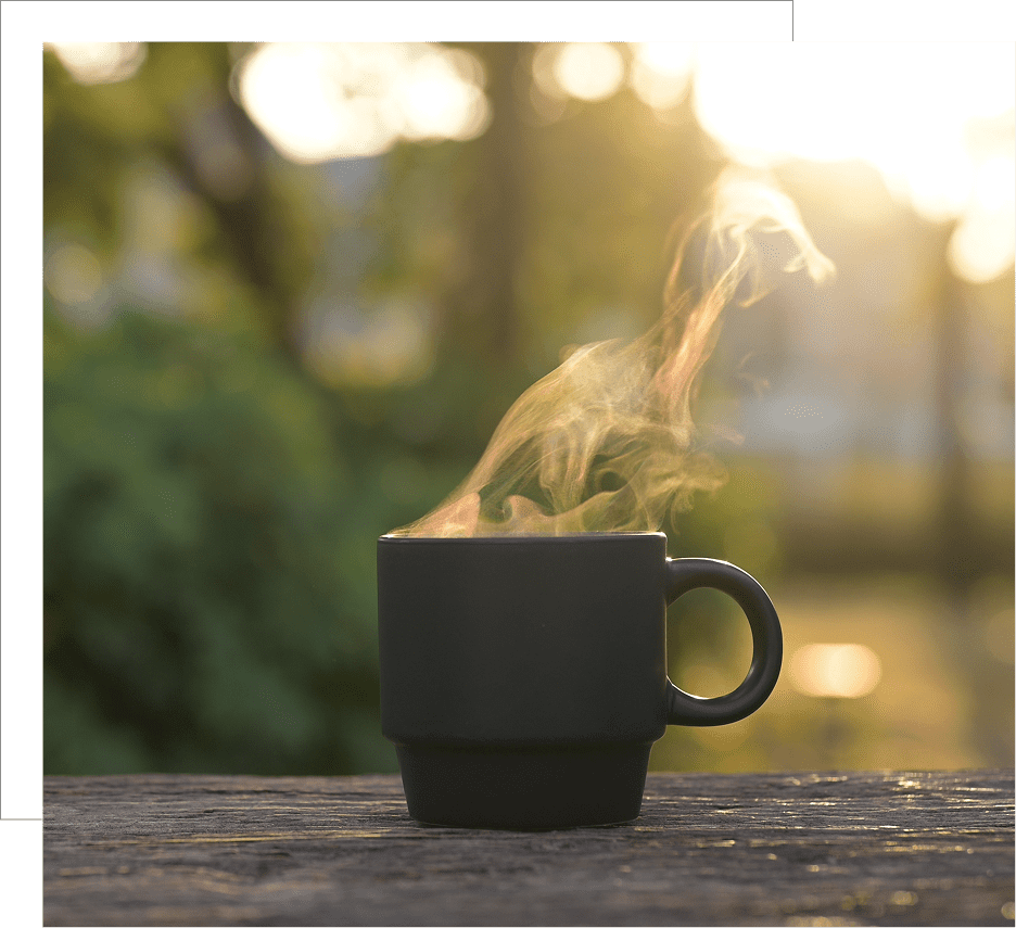 Steaming hot beverage in a black cup on a wooden table at sunrise.